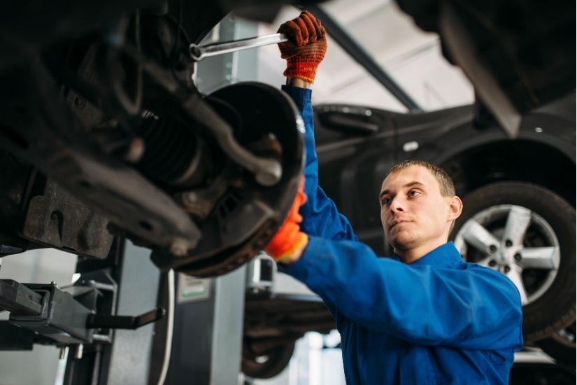 A mechanic with arm tattoos uses a tool to adjust or inspect the suspension strut and brake caliper inside the wheel well of a blue car.