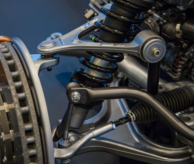 Close-up shot of a modern car's front suspension system featuring a silver upper control arm, coilover shock absorber, brake rotor, and steering rack components.