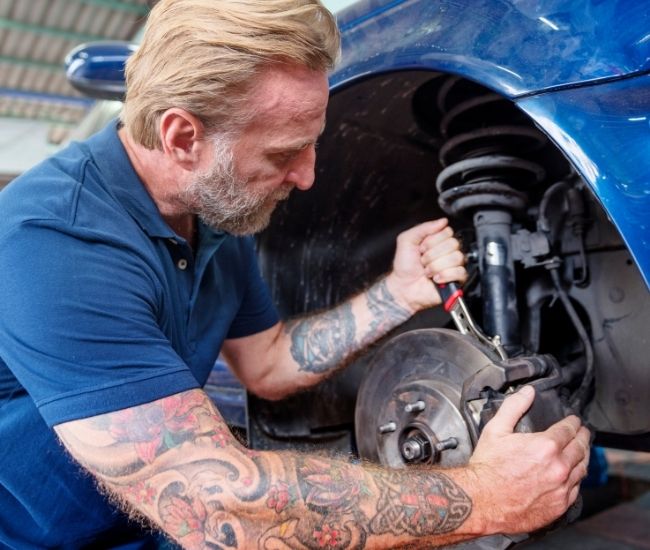 A mechanic with arm tattoos uses a tool to adjust or inspect the suspension strut and brake caliper inside the wheel well of a blue car.
