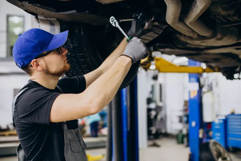 A mechanic in blue coveralls uses a wrench to tighten bolts on the underside of a vehicle that is raised on a professional workshop lift.