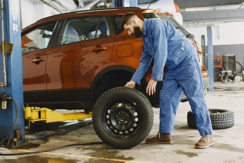 Professional mechanic in blue coveralls rolling a new tire toward an orange SUV in an automotive repair bay